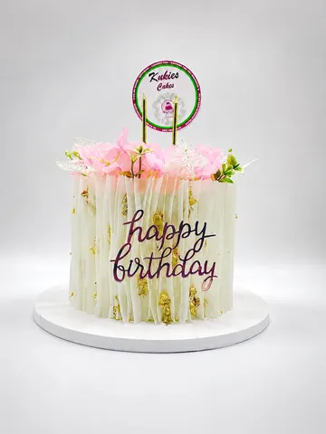 White cake with wafer-paper fringe texture, pink silk flowers and a gold Happy Birthday plaque