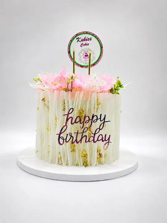 White cake with wafer-paper fringe texture, pink silk flowers and a gold Happy Birthday plaque