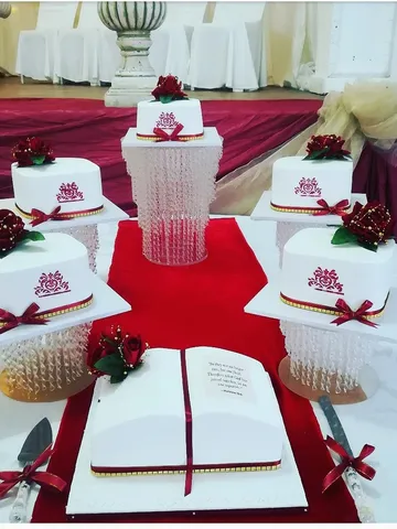 Wedding cake table with five white cakes, a maroon crest and red roses on crystal stands